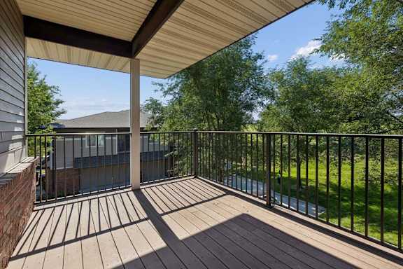 Covered apartment balcony with wood-look decking, black metal railings, and views of mature trees and landscaped green space.