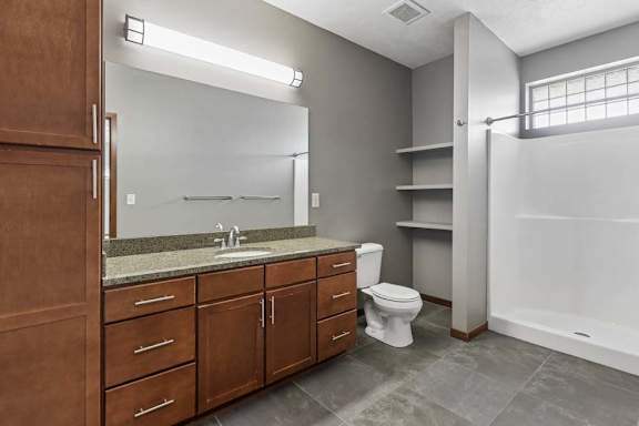 Apartment bathroom with wood cabinetry, stone-look vanity counter, open shelving, gray tile flooring, and a walk-in shower.