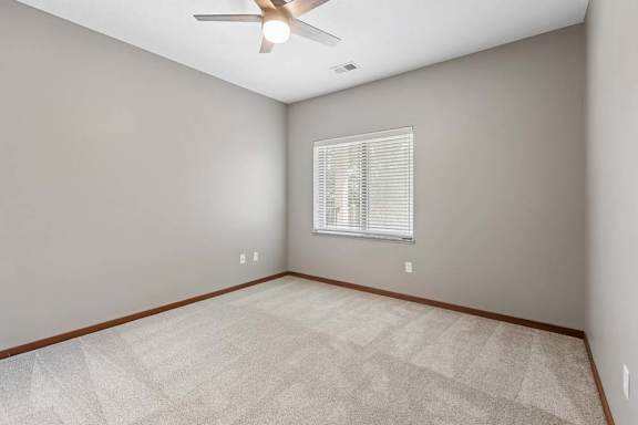 Bright, empty apartment bedroom with neutral carpet, gray walls, a ceiling fan, and a large window with blinds.
