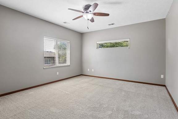 Bright, empty apartment bedroom with neutral carpet, gray walls, ceiling fan, and two windows with blinds.
