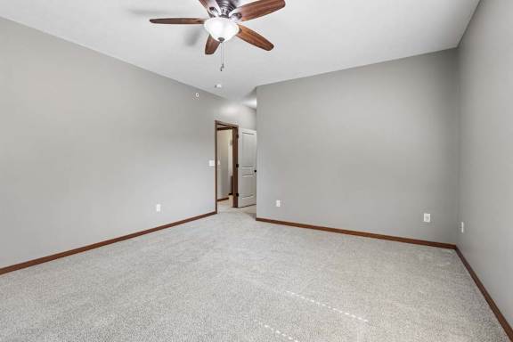 Spacious, empty apartment bedroom with neutral carpet, gray walls, wood trim, and a ceiling fan with light.