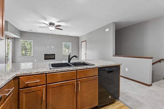 Granite kitchen peninsula with double sink overlooking a bright living space with fireplace, ceiling fan, and stairway access.