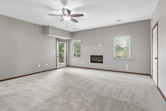 Bright, empty apartment living room with carpet, ceiling fan, two windows, a fireplace, and a glass door to the balcony.