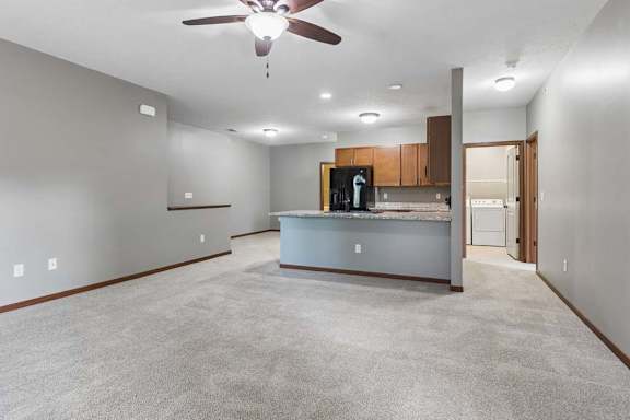 Open-concept apartment living area with carpet, ceiling fan, breakfast bar, wood cabinetry, black refrigerator, and laundry room access.