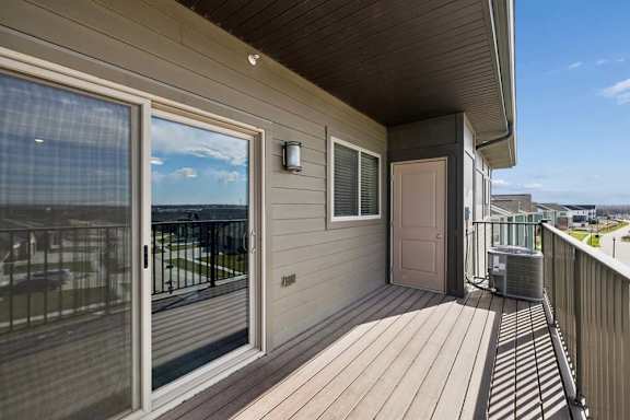 Covered apartment balcony with wood-style decking, a large sliding glass door, exterior lighting, and metal railings overlooking the surrounding community.