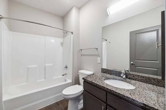 Apartment bathroom with a granite-look single-sink vanity, dark cabinetry, a large mirror, and a tub-shower combination with a white surround and modern fixtures.