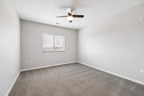 Spacious apartment bedroom with carpeted flooring, a ceiling fan, neutral walls, and a window with blinds providing natural light.