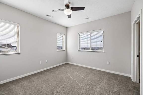 Spacious apartment bedroom with carpeted flooring, a ceiling fan, two windows with blinds providing natural light, and a sliding closet door.