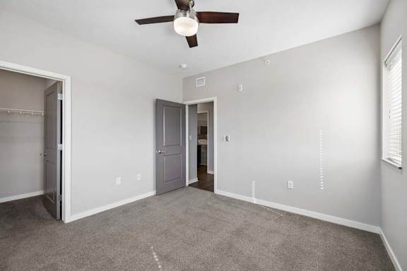 Spacious apartment bedroom with carpeted flooring, a ceiling fan, a walk-in closet, and a window with blinds providing natural light.