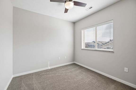 Bright apartment bedroom with carpeted flooring, a ceiling fan, neutral walls, and a window with blinds providing natural light.