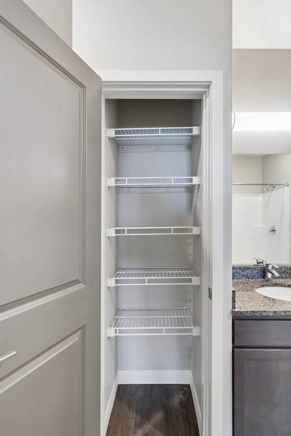 Apartment linen closet with built-in wire shelving, wood-style flooring, and double doors, located beside the bathroom vanity for convenient storage.