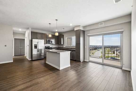 Spacious open-concept apartment living area with wood-style flooring, a modern kitchen featuring a center island, pendant lighting, stainless steel appliances, and a large sliding glass door leading to a private balcony.