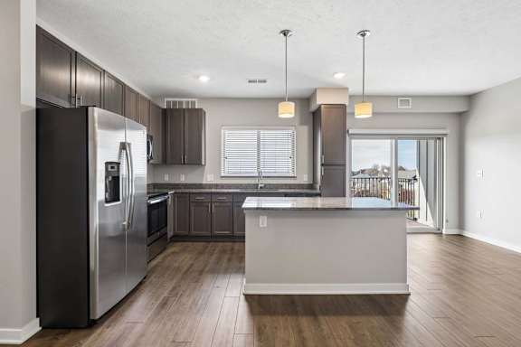 Open-concept apartment kitchen with a large island, dark cabinetry, granite-look countertops, stainless steel appliances, pendant lighting, and a spacious living area with a sliding glass door leading to a private balcony.