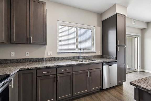 Modern apartment kitchen with dark cabinetry, granite-look countertops, a stainless steel dishwasher, and a large window above the sink providing natural light.