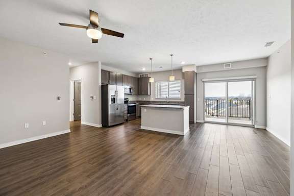 Spacious open-concept apartment living room with wood-style flooring, a ceiling fan, and a modern kitchen featuring a breakfast bar, dark cabinetry, stainless steel appliances, and a large sliding glass door leading to a private balcony.
