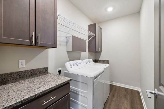 Apartment laundry room with side-by-side washer and dryer, granite-look countertop, dark cabinetry, built-in wire shelving, and wood-style flooring.