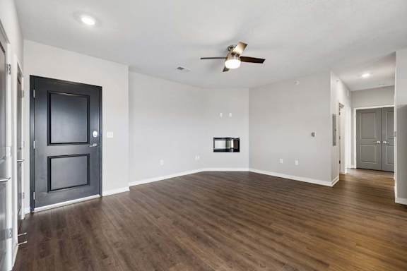 Spacious apartment living room with wood-style flooring, a ceiling fan, a wall-mounted fireplace, and an open layout with hallway access and a dark entry door.