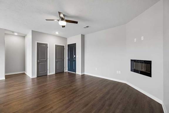 Spacious apartment living room with wood-style flooring, a ceiling fan, a wall-mounted fireplace, and an open layout with multiple interior doors and abundant natural light.