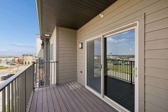 Covered apartment balcony with wood-style decking, metal railings, and a large sliding glass door overlooking the surrounding community.