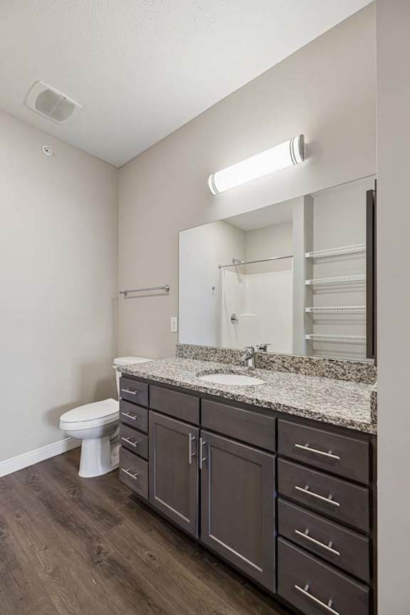 Apartment bathroom with a granite-look single-sink vanity, dark cabinetry, a large mirror with overhead lighting, and wood-style flooring.