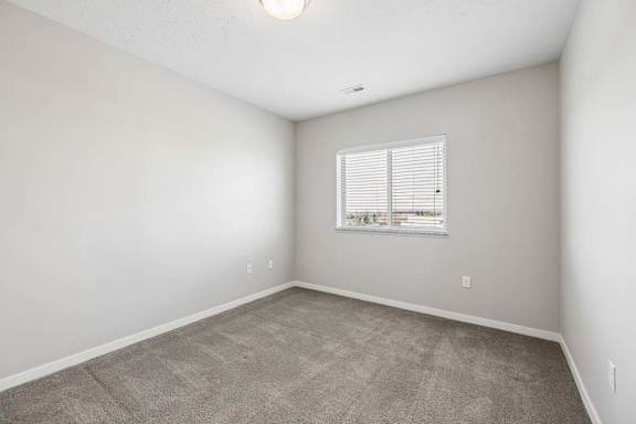 Bright apartment bedroom with carpeted flooring, neutral walls, and a window with blinds providing natural light.