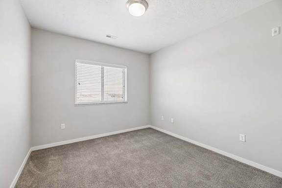 Bright apartment bedroom with carpeted flooring, neutral walls, and a window with blinds that provides natural light.