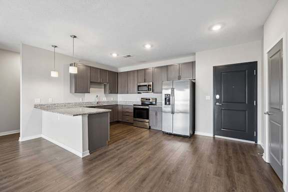 Open-concept apartment kitchen with a large island, dark cabinetry, granite-look countertops, stainless steel appliances, pendant lighting, and wood-style flooring.