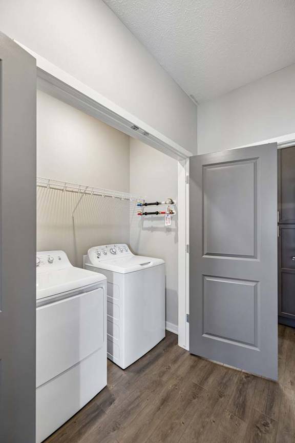 Apartment laundry room with side-by-side washer and dryer, built-in wire shelving, wood-style flooring, and a gray interior door leading to the hallway.