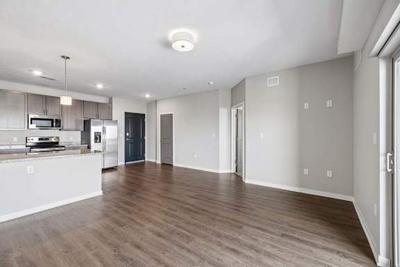 Spacious open-concept apartment living room with wood-style flooring, abundant natural light, and a modern kitchen featuring a breakfast bar, dark cabinetry, and stainless steel appliances.