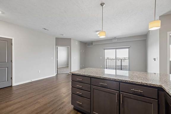 Open-concept apartment living area viewed from the kitchen island, featuring wood-style flooring, pendant lighting, and a large sliding glass door leading to a private balcony.