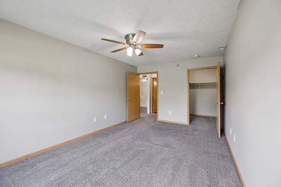 Spacious apartment bedroom with soft carpeting, a ceiling fan, an open closet, and access to an adjoining hallway.
