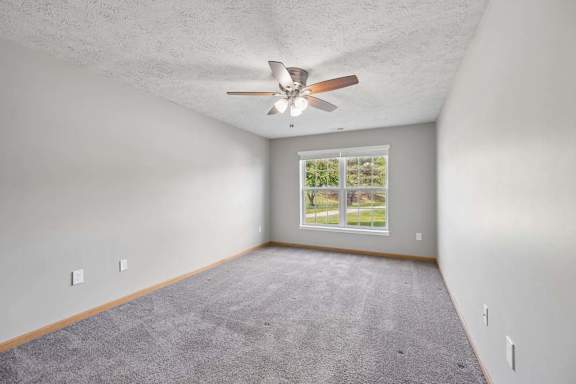 Bright apartment bedroom with soft carpeting, a ceiling fan, and a large window providing natural light.