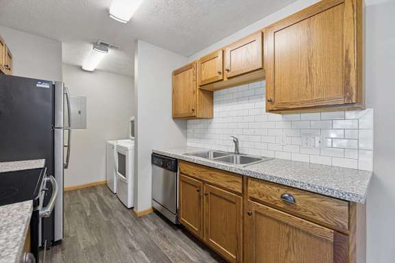 Galley-style apartment kitchen with wood cabinetry, a subway tile backsplash, stainless steel appliances, and an adjacent in-home laundry room.