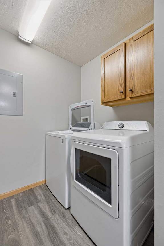 Dedicated apartment laundry room with a side-by-side washer and dryer, overhead storage cabinets, and wood-style flooring.