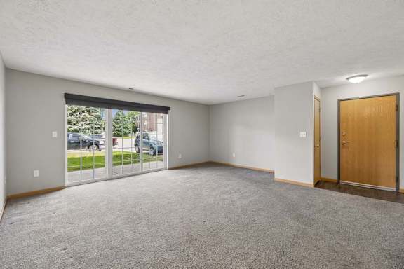 Spacious apartment living room with carpeted flooring, a large sliding glass door, and an open entryway with natural light.