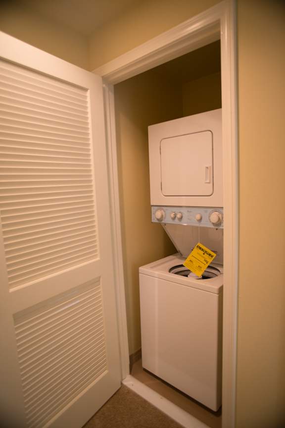 A small white washing machine is in a narrow space between a door with blinds and a wall.