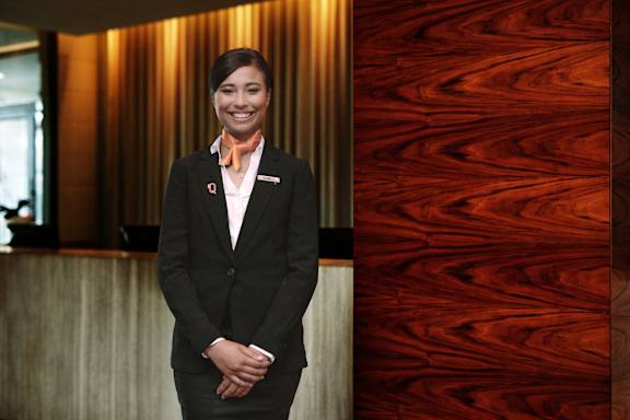 A woman in a black suit and a white shirt with a red tie is standing in front of a wooden wall.
