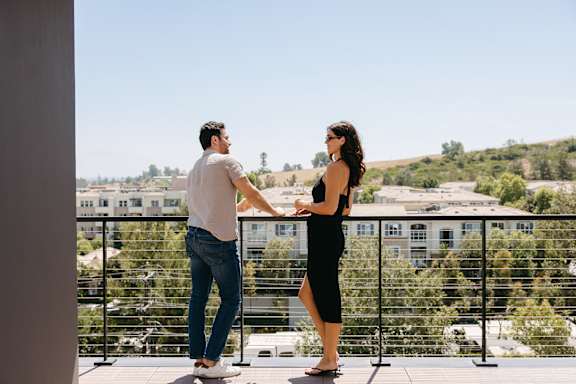 A man and a woman are standing on a balcony, holding hands.