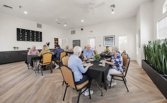 A group of elderly people are sitting around a table in a room with wooden floors and a blackboard on the wall.