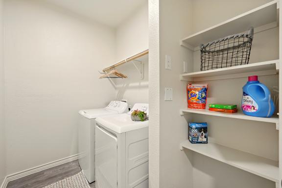 A white laundry room with a washer and dryer and a white shelving unit with cleaning supplies.