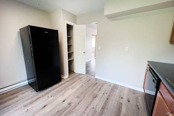A black fridge stands in a kitchen with a wooden floor and white walls.