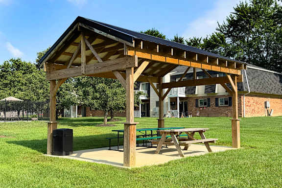 A wooden pavilion with a black roof is situated in a grassy area with a picnic table and a trash can.