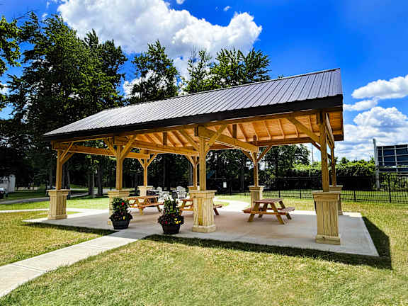 A wooden pavilion with a black roof and four pillars is surrounded by grass and trees.