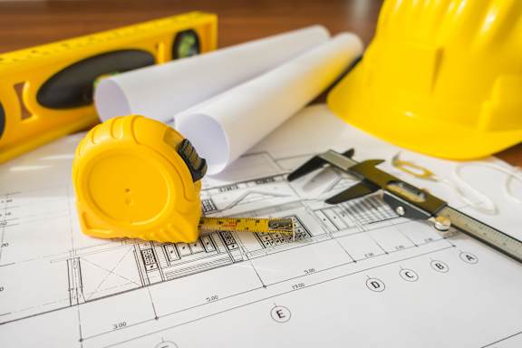 A yellow hard hat, a level, a pair of pliers, and rolled-up blueprints are on a wooden surface.