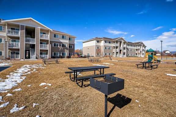 A picnic table is in the foreground of a grassy area with apartment buildings in the background.