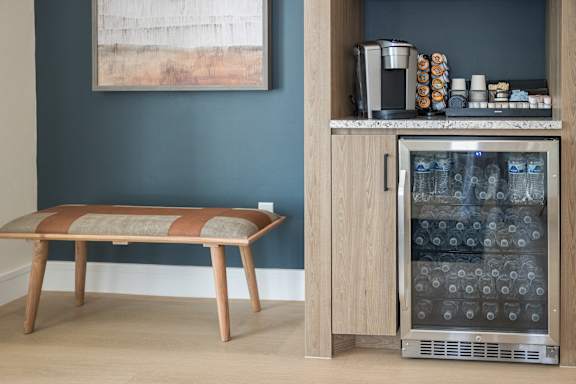 A wooden bench with a cushion sits in front of a wall with a painting and a wine cooler.