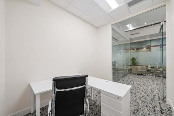 A black chair sits in front of a white desk in a room with a glass wall.