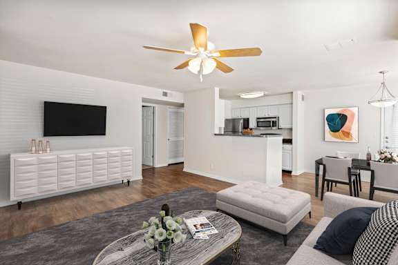 A modern living room with a flat screen TV, a coffee table with a vase of flowers, and a kitchen area in the background.