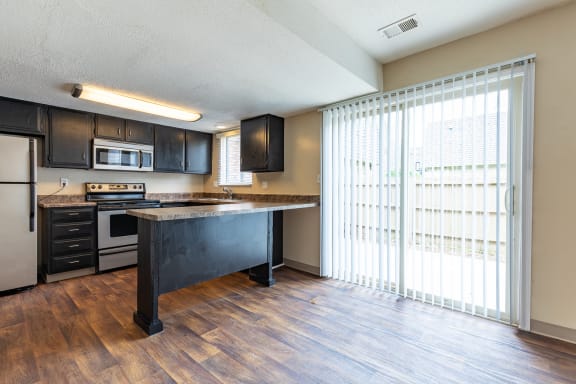 A kitchen with stainless steel appliances, hardwood floors, black storage cabinets, a bar stool seating area, and a sliding glass door patio entrance with vertical blinds.