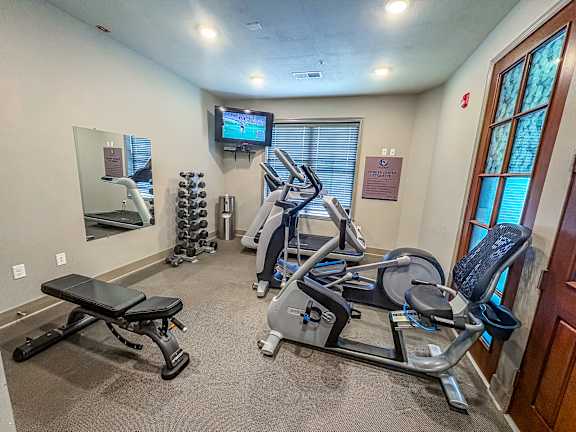Compact fitness center at The Dorset Apartments featuring cardio machines, weight bench, dumbbells, and a mounted TV.
